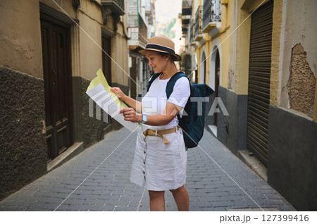 Woman Tourist Exploring a Scenic Medieval City Street with a Map. Woman tourist in summer attire and a backpack explores a charming historic street while reading a map. 127399416
