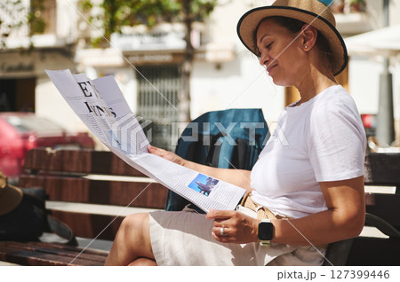 Smiling Woman Reading a Newspaper Outdoors in a Relaxed Urban Setting 127399446