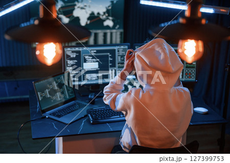 Sitting in white hoodie. Young professional female hacker is indoors by computer with lot of information on displays 127399753
