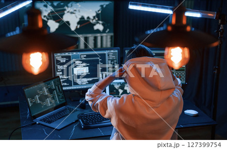 Sitting in white hoodie. Young professional female hacker is indoors by computer with lot of information on displays 127399754
