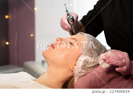 Aesthetician performs facial microdermabrasion with airbrush and cotton pad 127400034