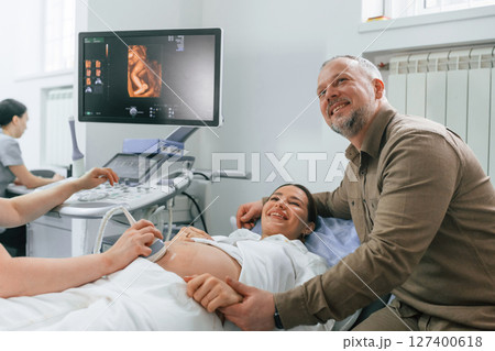 Husband is supporting his wife. Pregnant woman is lying down in the hospital, doctor does ultrasound Husband is supporting his wife. Pregnant woman is lying down in the hospital, doctor does ultrasound 127400618