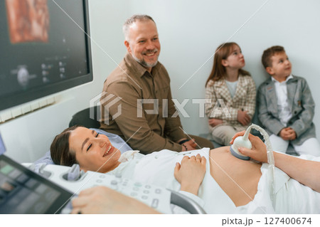 Husband and kids are sitting together. Pregnant woman is lying down in the hospital, doctor does ultrasound 127400674
