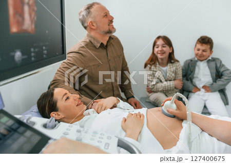 Husband and kids are sitting together. Pregnant woman is lying down in the hospital, doctor does ultrasound 127400675