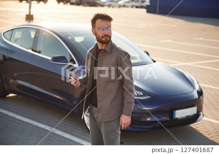 Holding smartphone in hand. Man is standing near his electric car outdoors 127401687