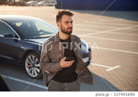 Holding smartphone in hand. Man is standing near his electric car outdoors 127401699