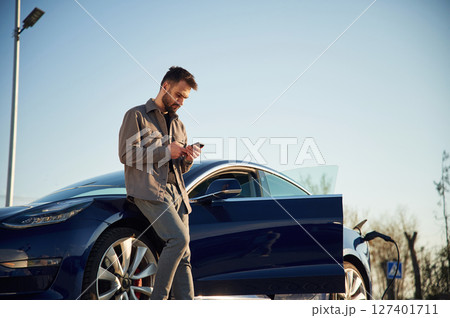 Charging the automobile. Man is standing near his electric car outdoors Charging the automobile. Man is standing near his electric car outdoors 127401711