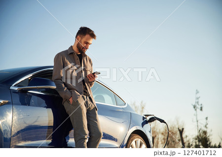 Holding smartphone in hand. Man is standing near his electric car outdoors 127401712