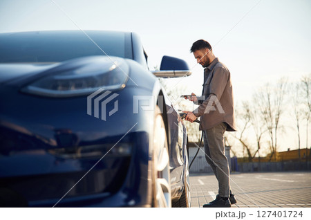 View from the side. Charging the automobile. Man is standing near his electric car outdoors View from the side. Charging the automobile. Man is standing near his electric car outdoors 127401724