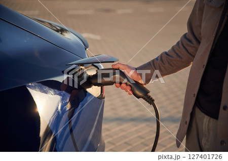 Blue colored automobile. Putting the charger in. Man is standing near his electric car outdoors 127401726