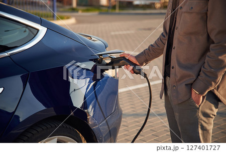 Blue colored automobile. Putting the charger in. Man is standing near his electric car outdoors Blue colored automobile. Putting the charger in. Man is standing near his electric car outdoors 127401727
