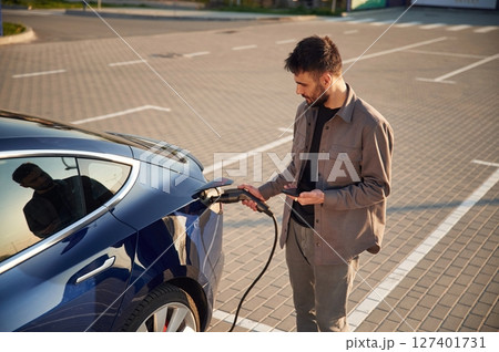 Holding the charger. Man is standing near his electric car outdoors Holding the charger. Man is standing near his electric car outdoors 127401731