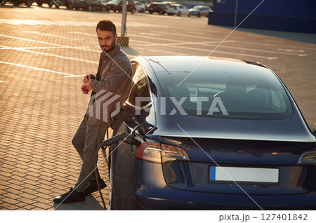 On the charging station. Man is standing near his electric car outdoors On the charging station. Man is standing near his electric car outdoors 127401842