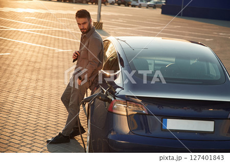 On the charging station. Man is standing near his electric car outdoors On the charging station. Man is standing near his electric car outdoors 127401843