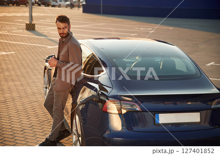 Charging process. Man is standing near his electric car outdoors Charging process. Man is standing near his electric car outdoors 127401852