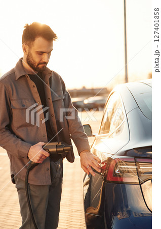On the parking lot. Charging the automobile. Man is standing near his electric car outdoors 127401858