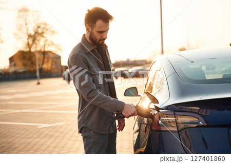 On the parking lot. Charging the automobile. Man is standing near his electric car outdoors 127401860