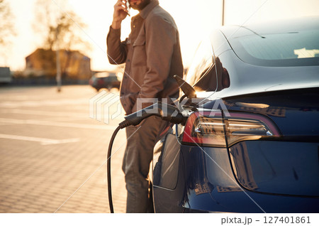 Talking by phone. Man is standing near his electric car outdoors Talking by phone. Man is standing near his electric car outdoors 127401861