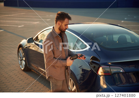 Putting charger into socket. Man is standing near his electric car outdoors Putting charger into socket. Man is standing near his electric car outdoors 127401880