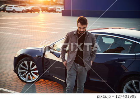 Sunny background. Man is standing near his electric car outdoors 127401908