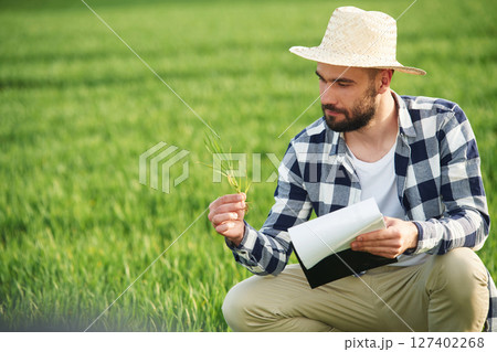 In straw hat. Holding wheat and notepad. Handsome young man is on agricultural field 127402268