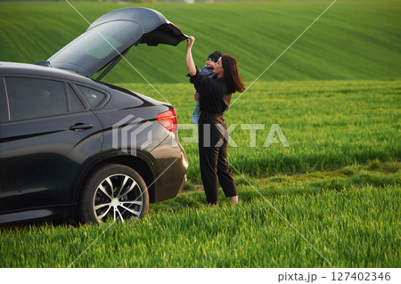 Standing near car, closing trunk. Mother with her little baby son is outdoors on the agricultural field 127402346