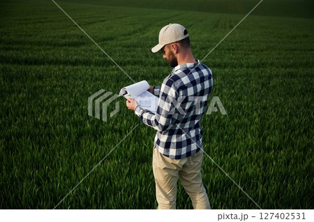Standing, holding document. In cap. Handsome young man is on agricultural field Standing, holding document. In cap. Handsome young man is on agricultural field 127402531