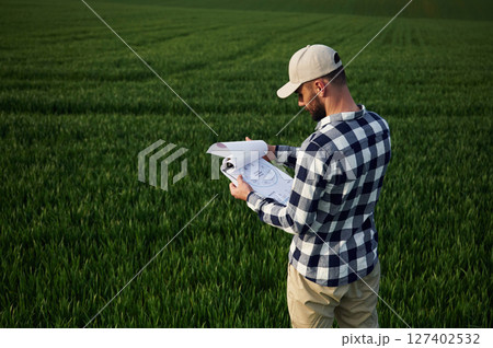 Standing, holding document. In cap. Handsome young man is on agricultural field Standing, holding document. In cap. Handsome young man is on agricultural field 127402532