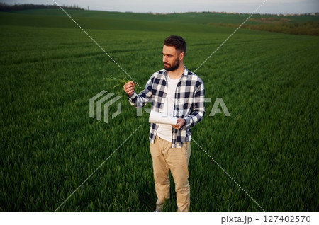Front view. Standing and holding notepad with document. Handsome young man is on agricultural field Front view. Standing and holding notepad with document. Handsome young man is on agricultural field 127402570