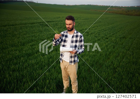 Front view. Standing and holding notepad with document. Handsome young man is on agricultural field Front view. Standing and holding notepad with document. Handsome young man is on agricultural field 127402571