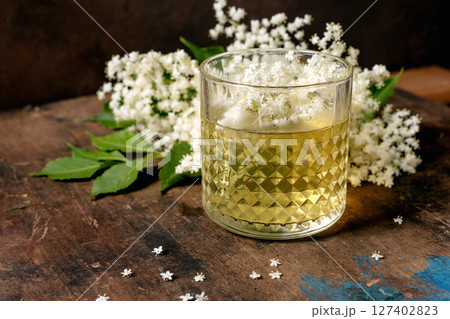 Crystal glass with elderflower cocktail decorated with white elderflower blossoms on rustic wooden table. Natural herbal drink with floral garnish. 127402823