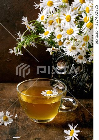 Glass cup herbal chamomile tea with white daisy flowers in vintage vase on wooden table. Natural wellness drink concept with fresh blooms decoration. 127402825