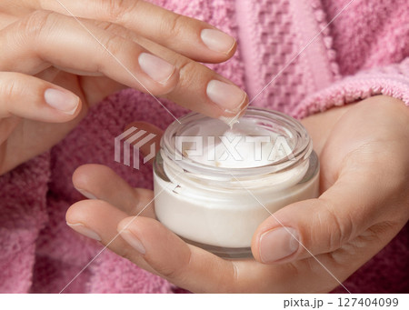 Young woman in pink bathrobe touching cream with finger and holding a cosmetic jar closeup 127404099