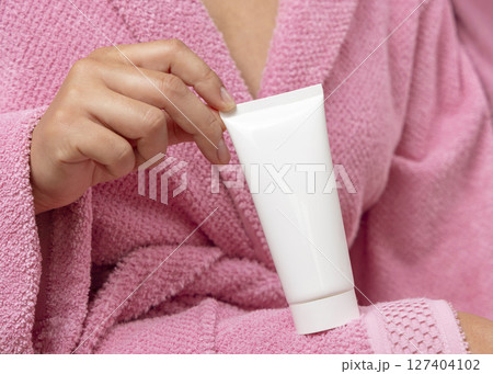 Closeup of young woman in pink bathrobe holding squeeze cream tube in hands, cosmetic mockup 127404102