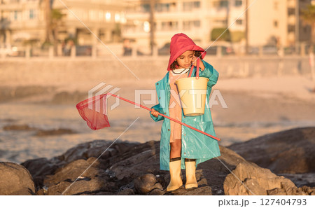 Beach, fishing and girl with a net and bucket standing on a rock by the ocean on vacation. Outdoor, nature and child at the seaside to catch fish in the sea water for an adventure while on holiday. 127404793