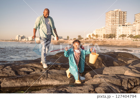 Father, girl and fishing at beach, learning and happy together outdoor in summer, bonding or fun on rocks. Black man teaching kid to catch fish, rod and bucket with family at a ocean, sea and nature 127404794