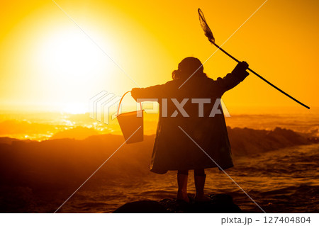 Silhouette, sunset and child at beach with toys for travel, holiday and playing by water in Australia. Earth day, ocean and back of girl kid in the dark during sunrise to play and relax on vacation 127404804