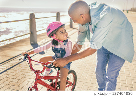 Father teaching his child to ride a bike at the beach while on a summer vacation, holiday or adventure. Happy, learning and man helping his girl kid with her bicycle while bonding in nature by ocean. 127404850