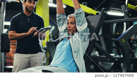 Personal trainer, senior woman and machine at gym for chest press, muscle strength and wellness. Retirement fitness, instructor and tablet for workout progress, exercise monitor and training support 127407489