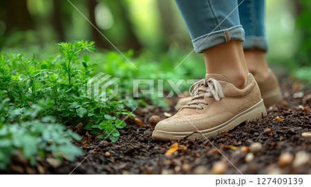 Person wearing beige sneakers stands on soil next to green plants in garden.  127409139