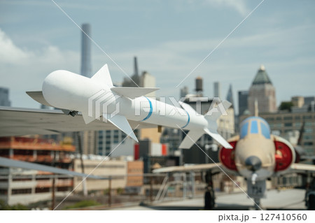 A white air-to-air missile is mounted under the wing of a military jet, photographed with an urban skyline in the background. The missile features stabilizing fins and blue rings. 127410560