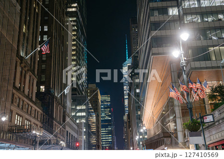 Multiple American flags line the street between tall Midtown skyscrapers glowing under the night sky. The scene is lit by streetlamps and reflects the patriotic spirit of New York City. 127410569