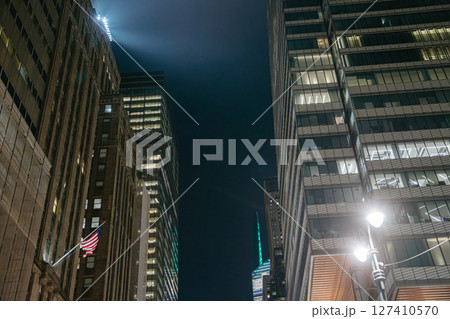 Tall buildings rise into the night sky in Midtown Manhattan, with an American flag hanging from one facade. Bright lights from street lamps and office windows illuminate the cityscape. 127410570