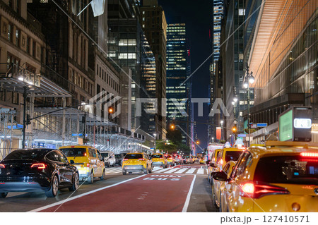 Yellow taxis and a black car drive through Midtown Manhattan at night under skyscrapers and streetlights. The scene features busy traffic, scaffolding, and the bright glow of the city. 127410571