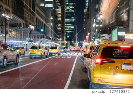 A row of yellow taxis waits in traffic on a Midtown Manhattan street at night. Skyscrapers, glowing lights, and urban movement create a classic New York atmosphere. 127410573