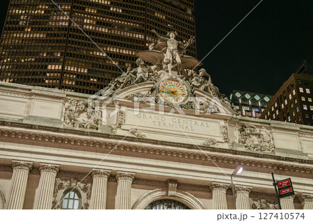 The famous Grand Central Terminal clock is illuminated above the main entrance, surrounded by classical statues. Tall skyscrapers tower behind the detailed architecture in Midtown Manhattan. 127410574