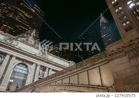 The illuminated facade of Grand Central Terminal glows against the New York City skyline at night. The iconic Chrysler Building rises in the background, adding to the historic cityscape. 127410575