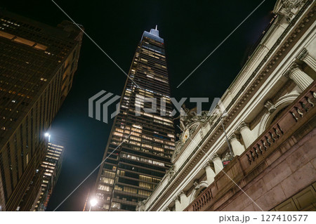 A tall modern skyscraper rises sharply into the night sky beside the historic facade of Grand Central Terminal in New York City. The illuminated windows and architectural contrast capture the dynamic 127410577