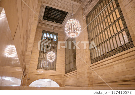 Two ornate chandeliers hang from the high ceiling of a stone corridor inside Grand Central Terminal. Tall arched windows with decorative ironwork surround the warm interior lighting. 127410578