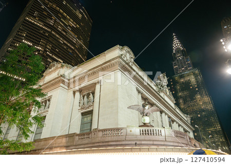 The iconic facade of Grand Central Terminal is lit up at night in New York City. Behind it rise tall modern skyscrapers, including the Chrysler Building, blending classic and modern architecture. The iconic facade of Grand Central Terminal is lit up at night in New York City. Behind it rise tall modern skyscrapers, including the Chrysler Building, blending classic and modern architecture. 127410594